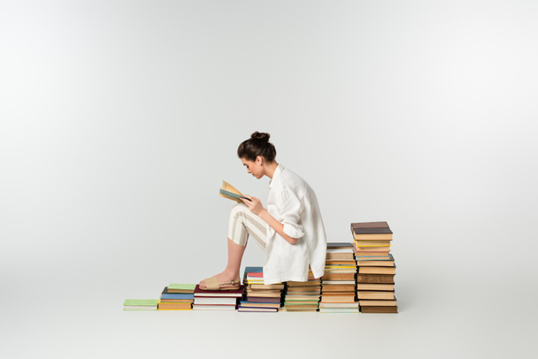 πλαϊνή άποψη της νεαρής γυναίκας σε σανδάλια ανάγνωση, ενώ κάθεται σε σωρό από βιβλία σχετικά με το λευκό  - Φωτογραφία, εικόνα
