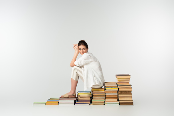 full length of smiling woman in sandals sitting on pile of books on white  - Photo, Image