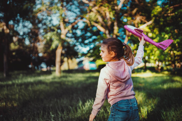 Little girl launches a toy plane into the air in the park outdoor. Child launches a toy plane. Beautiful little girl stands on the grass and launches a pink toy plane. - Фото, изображение