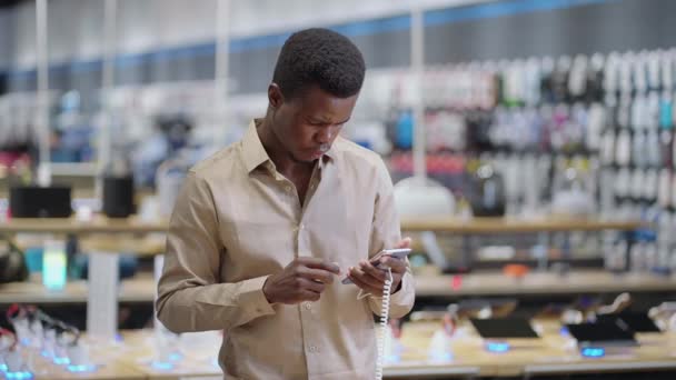 seriózní afro-americký muž je prohlížení smartphone v obchodě s digitálním vybavením, testování nových modelů před nákupem - Záběry, video