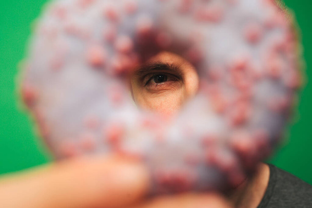 close-up jongeman dekt oog omhoog met donut, geïsoleerd op groene achtergrond - Foto, afbeelding