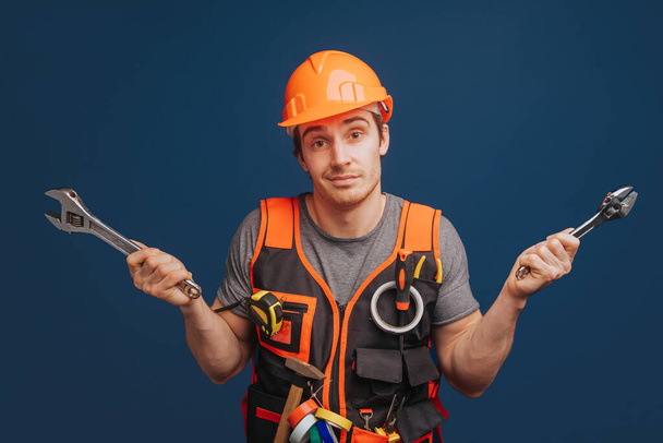 Bewildered builder in a helmet is holding wrenches, isolated over blue background - Photo, Image