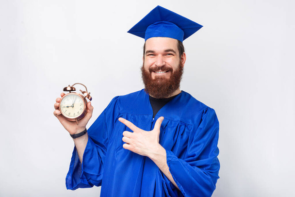 Улыбающийся бородатый выпускник указывает на будильник, который он держит, на белом фоне. - Фото, изображение