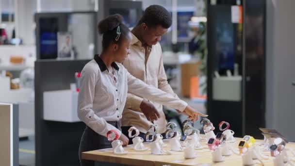 zwart paar in digitale apparatuur winkel, vrouwelijke en mannelijke shoppers bekijken modellen van smartwatch, bespreken monsters op demonstratie showcase - Video