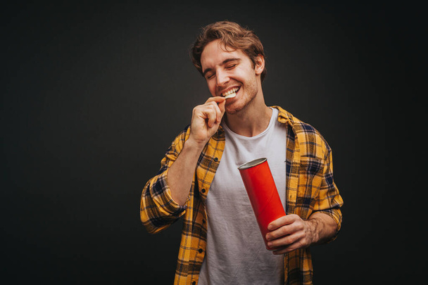 Joven hombre rubio en camisa amarilla está comiendo patatas fritas, aislado sobre fondo negro - Foto, Imagen