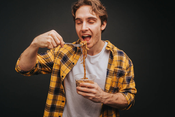 Joven hombre rubio con camisa amarilla sosteniendo tenedor de espagueti antes de comer, aislado sobre fondo negro - Foto, Imagen