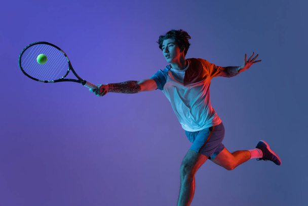 Young caucasian man playing tennis isolated on purple-blue studio background in neon, action and motion concept - Fotó, kép
