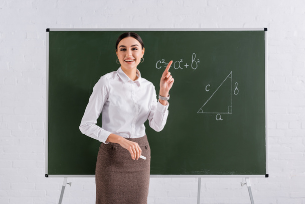Lachende leraar wijzend met vinger in de buurt van schoolbord met wiskundige formule  - Foto, afbeelding