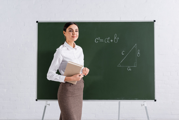 Brunette teacher with book and chalk smiling near equation on chalkboard  - Photo, Image
