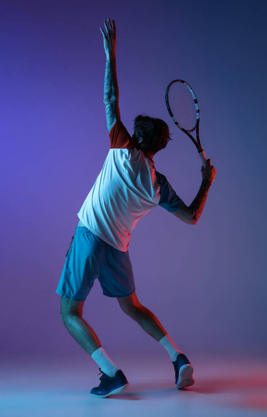Young caucasian man playing tennis isolated on purple-blue studio background in neon, action and motion concept - Фото, изображение