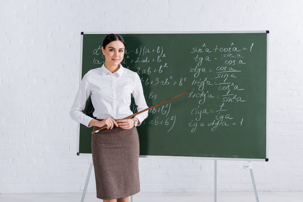 Teacher with pointer smiling at camera near chalkboard during mathematic lesson  - Photo, Image