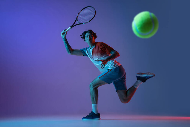 Jeune homme caucasien jouant au tennis isolé sur fond studio violet-bleu au néon, concept d'action et de mouvement - Photo, image