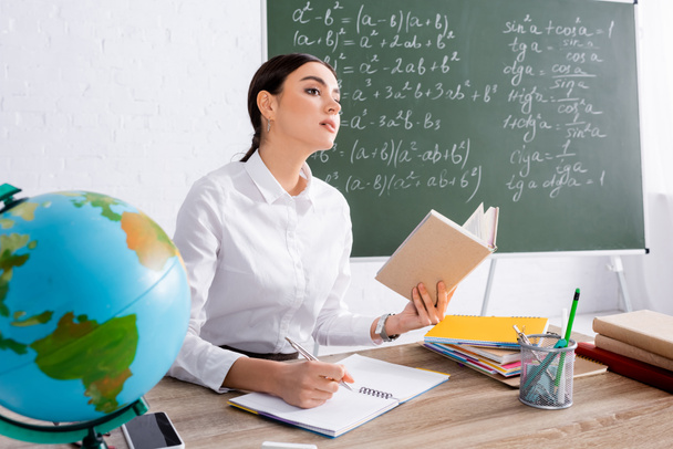 Teacher holding book and writing on notebook near globe in classroom  - Foto, imagen