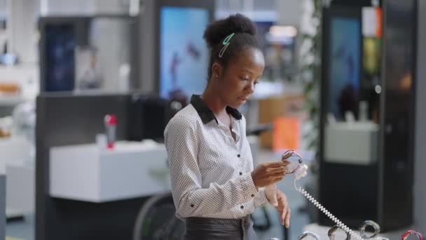 mujer negra en la tienda de equipos digitales, el comprador femenino está eligiendo un nuevo modelo de reloj inteligente, viendo la muestra en el escaparate de demostración - Metraje, vídeo