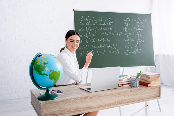 Uśmiechnięty nauczyciel wskazujący tablicę podczas lekcji online na laptopie w pobliżu globu i książek  - Zdjęcie, obraz