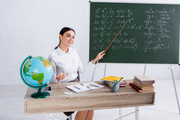 Χαρούμενος δάσκαλος με δείκτη κάθεται κοντά στο chalkboard και υδρόγειο κατά τη διάρκεια του μαθήματος  - Φωτογραφία, εικόνα