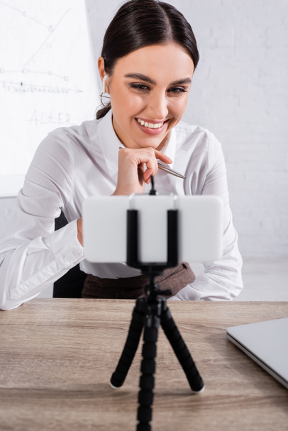 Cheerful businesswoman in earphone having video chat on blurred smartphone in office  - Fotó, kép