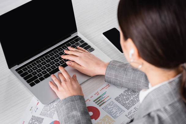 Verschwommene Geschäftsfrau benutzt Laptop mit leerem Bildschirm in der Nähe von Zeitungen mit Diagrammen  - Foto, Bild
