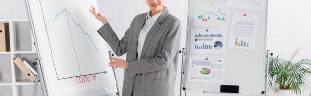 Обрезанный вид улыбающейся деловой женщины, указывающей на флипчарт с надписью covid-2019, баннер  - Фото, изображение