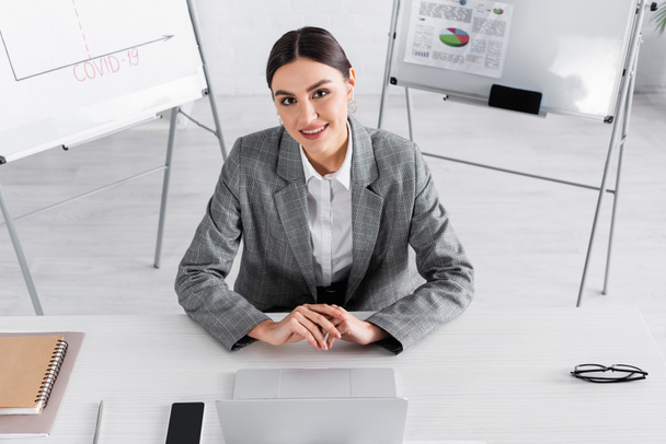 Высокий угол обзора деловой женщины, улыбающейся в камеру рядом с устройствами на рабочем столе  - Фото, изображение
