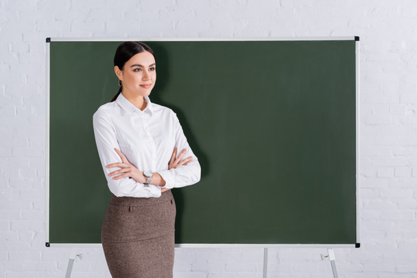 Young teacher with crossed arms looking away near chalkboard  - Φωτογραφία, εικόνα