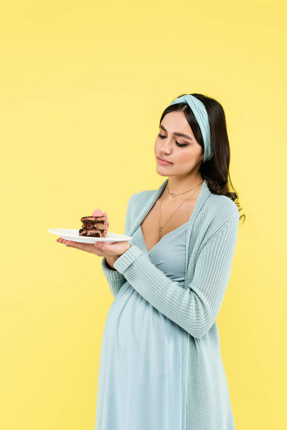 молодая беременная женщина испытывает сомнения, держа вкусный торт изолирован на желтый - Фото, изображение