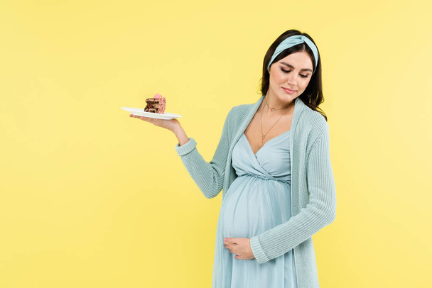 pregnant woman touching belly while holding piece of tasty cake isolated on yellow - Foto, imagen