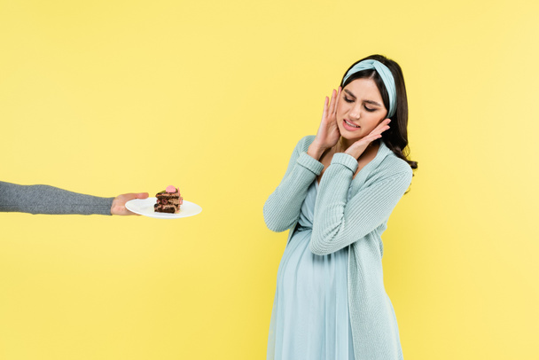 беременная женщина чувствует отвращение рядом с кусочком вкусного торта изолированы на желтый - Фото, изображение