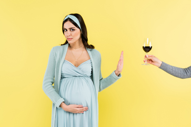 беременная женщина показывает стоп-жест рядом с бокалом вина изолированы на желтый - Фото, изображение