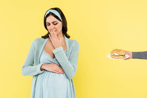 niezadowolona ciężarna kobieta zakrywająca usta dłonią w pobliżu hamburgera izolowanego na żółto - Zdjęcie, obraz