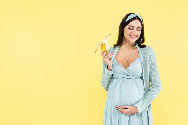usmívající se těhotná žena drží zralé banány izolované na žluté - Fotografie, Obrázek