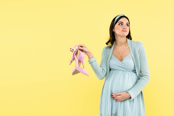 στοχαστική έγκυος γυναίκα που κρατά ψηλά τακούνια παπούτσια απομονώνονται σε κίτρινο - Φωτογραφία, εικόνα