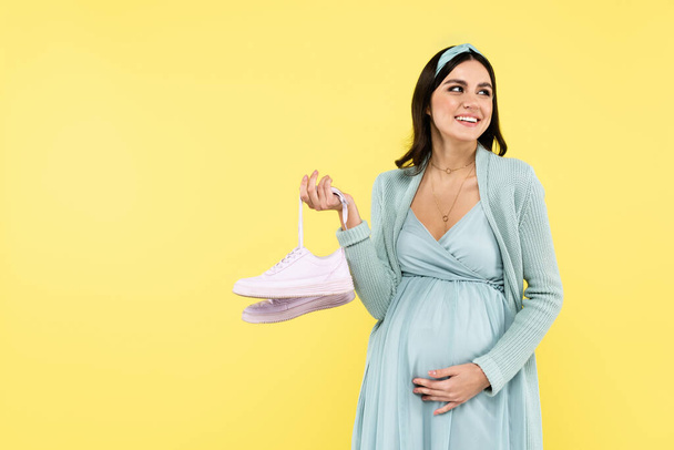 pleased pregnant woman touching belly while holding sneakers isolated on yellow - Foto, Bild