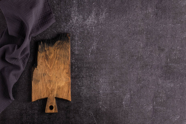 An old dark cutting board on a dark background. Cutting board for food. The board is charred. Signature background. Grey linen napkin - Zdjęcie, obraz