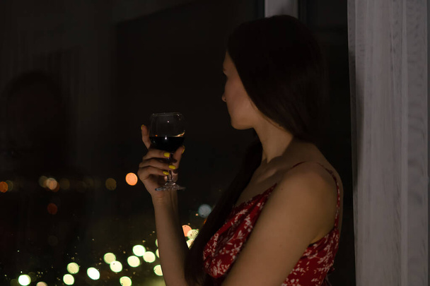 Jeune femme brune en robe rouge avec des bretelles et un verre de vin rouge se tient près de la fenêtre et regarde la vie nocturne de la ville. Focus sélectif - Photo, image