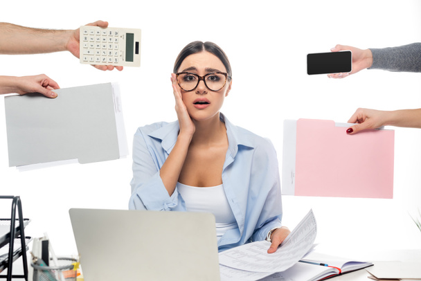 shocked businesswoman looking at camera near devices and coworkers with documents isolated on white - Photo, image