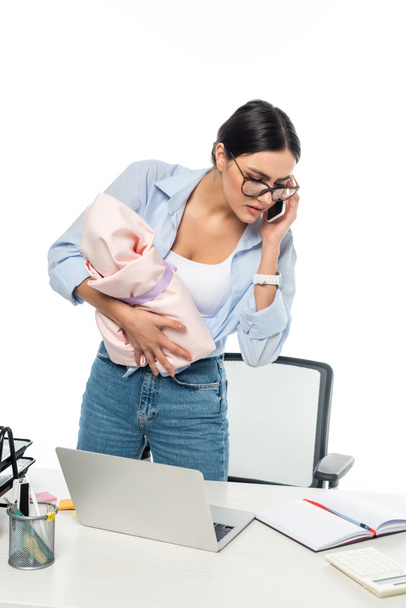 νεαρή μητέρα μιλάει στο κινητό ενώ κρατά το μωρό κοντά στο γραφείο εργασίας απομονωμένο στα λευκά - Φωτογραφία, εικόνα