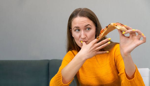 Mulher de cabelos escuros bonita jovem em uma camisola laranja está segurando um pedaço de pizza em uma mão, e lambendo o dedo da outra mão. Concentra-te na mulher. Banner - Foto, Imagem