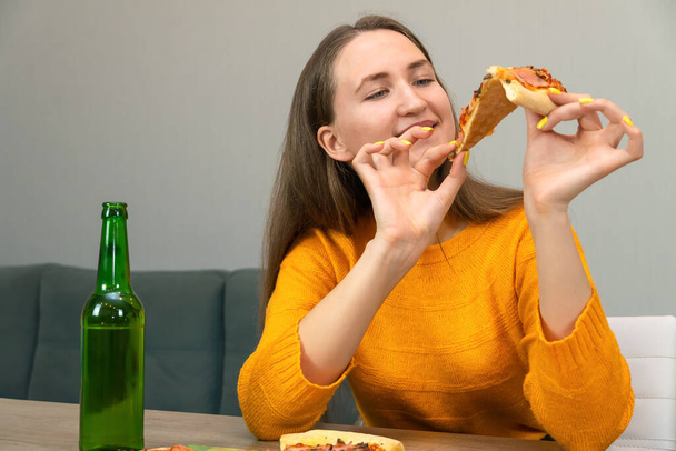 Hermosa joven morena con una sudadera naranja sostiene una rebanada de pizza con ambas manos y sonrisas, junto a una cerveza en una botella verde - Foto, Imagen
