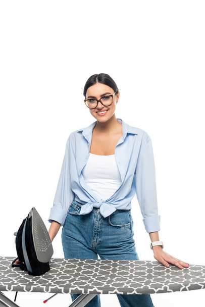 young woman in eyeglasses smiling at camera near iron and ironing board isolated on white - Foto, afbeelding