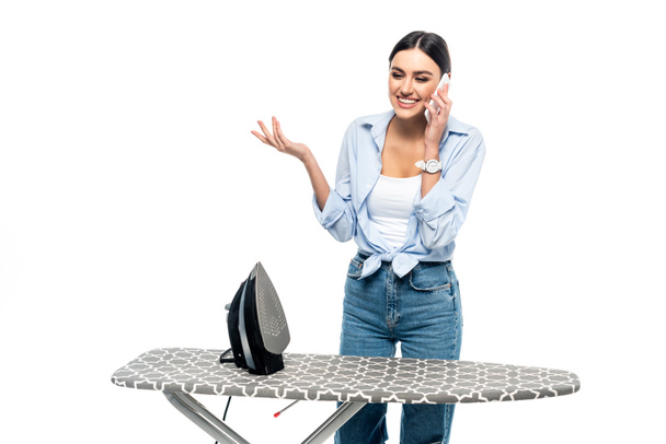 cheerful woman gesturing while talking on cellphone near ironing board isolated on white - 写真・画像