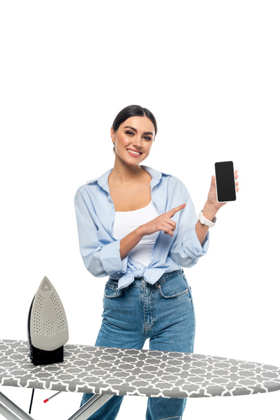 happy housewife pointing at smartphone with blank screen near ironing board isolated on white - Photo, image