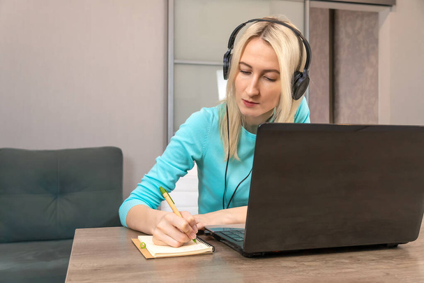 Young blonde woman sitting at a desk with large headphones at a laptop holding a pen, writing notes in a notebook,listening to a lecture or webinar, learning via the Internet,improving language skills - Photo, Image