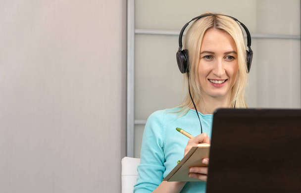 Νεαρή χαμογελαστή ξανθιά γυναίκα κάθεται σε ένα γραφείο με μεγάλα ακουστικά σε ένα φορητό υπολογιστή, κρατά ένα στυλό και γράφει σημειώσεις σε ένα σημειωματάριο, ακούει μια διάλεξη ή webinar, μαθαίνει μέσω του Διαδικτύου, βελτιώνει τις γλωσσικές δεξιότητες - Φωτογραφία, εικόνα