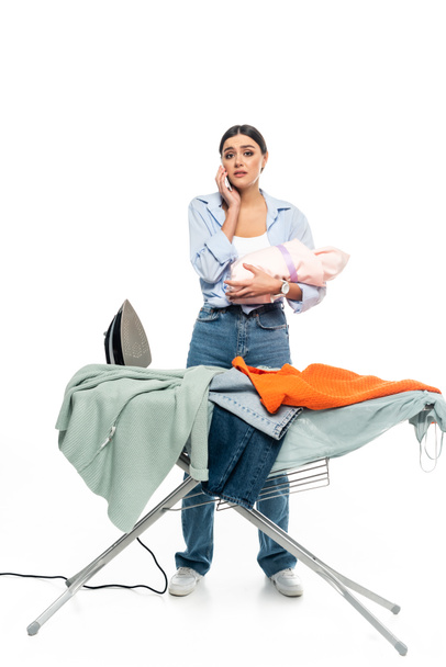 mujer cansada con bebé bebé hablando en el teléfono inteligente cerca de tabla de planchar sobre fondo blanco - Foto, imagen