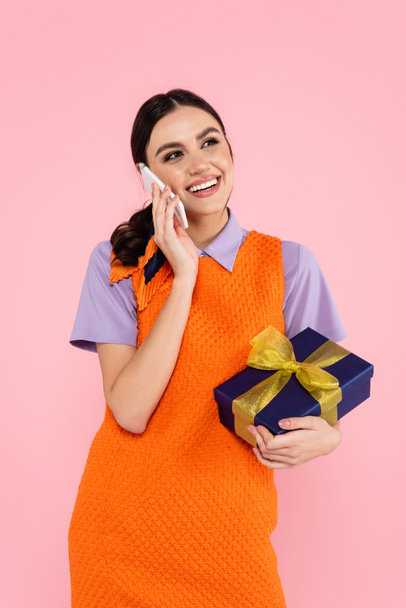 happy woman holding gift box while talking on smartphone isolated on pink - Zdjęcie, obraz