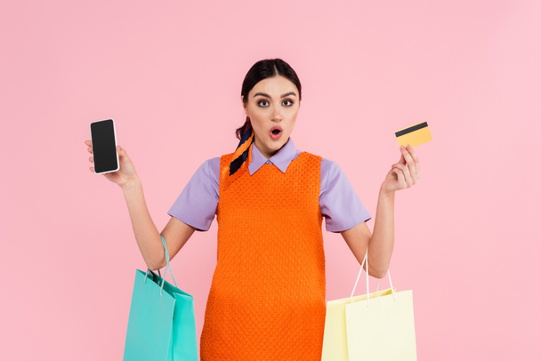 amazed woman showing smartphone and credit card while holding shopping bags isolated on pink - Foto, Imagem