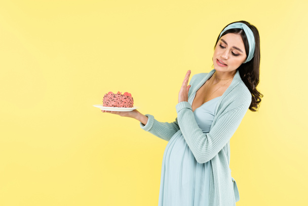 έγκυος γυναίκα δείχνει άρνηση χειρονομία κρατώντας γλυκό επιδόρπιο απομονώνονται σε κίτρινο - Φωτογραφία, εικόνα