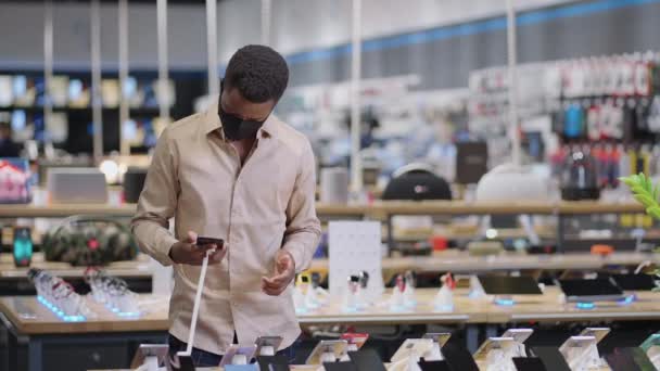 μαύρος άνδρας με μάσκα προσώπου δοκιμάζει smartphone σε κατάστημα ψηφιακού εξοπλισμού, προβολή νέων μοντέλων πριν από την αγορά, πορτρέτο του αρσενικού αγοραστή - Πλάνα, βίντεο