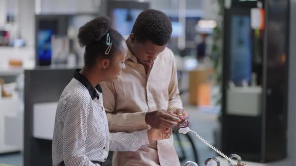 mężczyzna i kobieta robią zakupy w sklepie z cyfrowym sprzętem, czarna para przegląda nowy model smartwatcha, pobierając próbki z gabloty pokazowej - Materiał filmowy, wideo